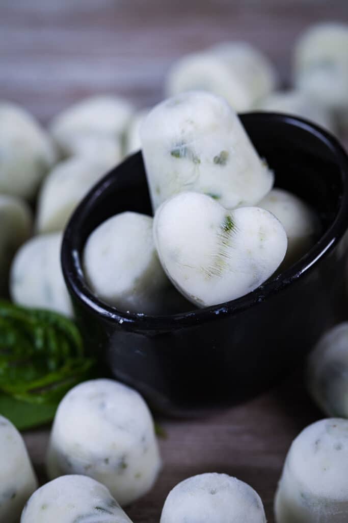 A close up of white ice cubes in a small bowl and surrounded by randomly placed ice cubes on a wooden board.