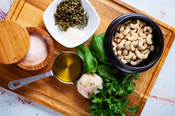 Ingredients like salt, capers, olive oil, garlic, cashews, parsley, basil leaves, dried Rosemary needles, and onion powder presented on a wooden board.