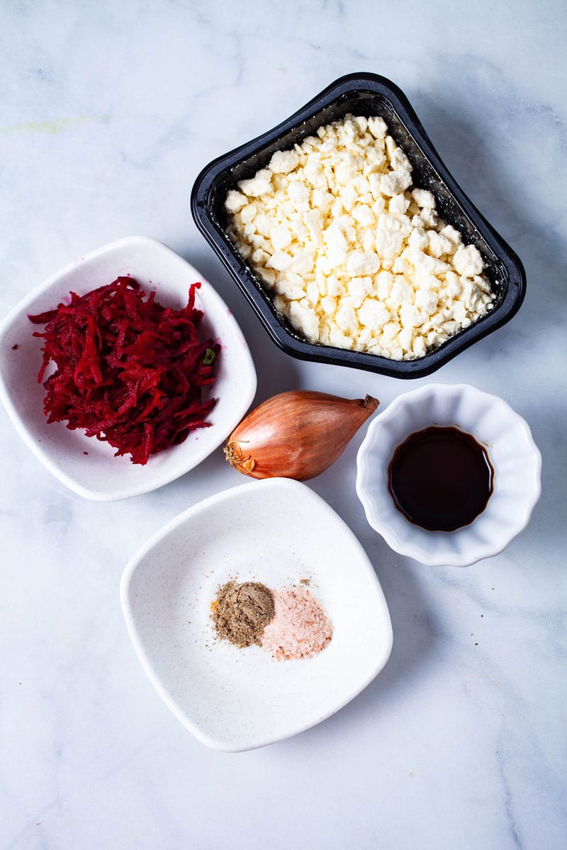 Grated beets, a shallot, feta cheese, balsamic vinegar, and a set of spices displayed on a table.