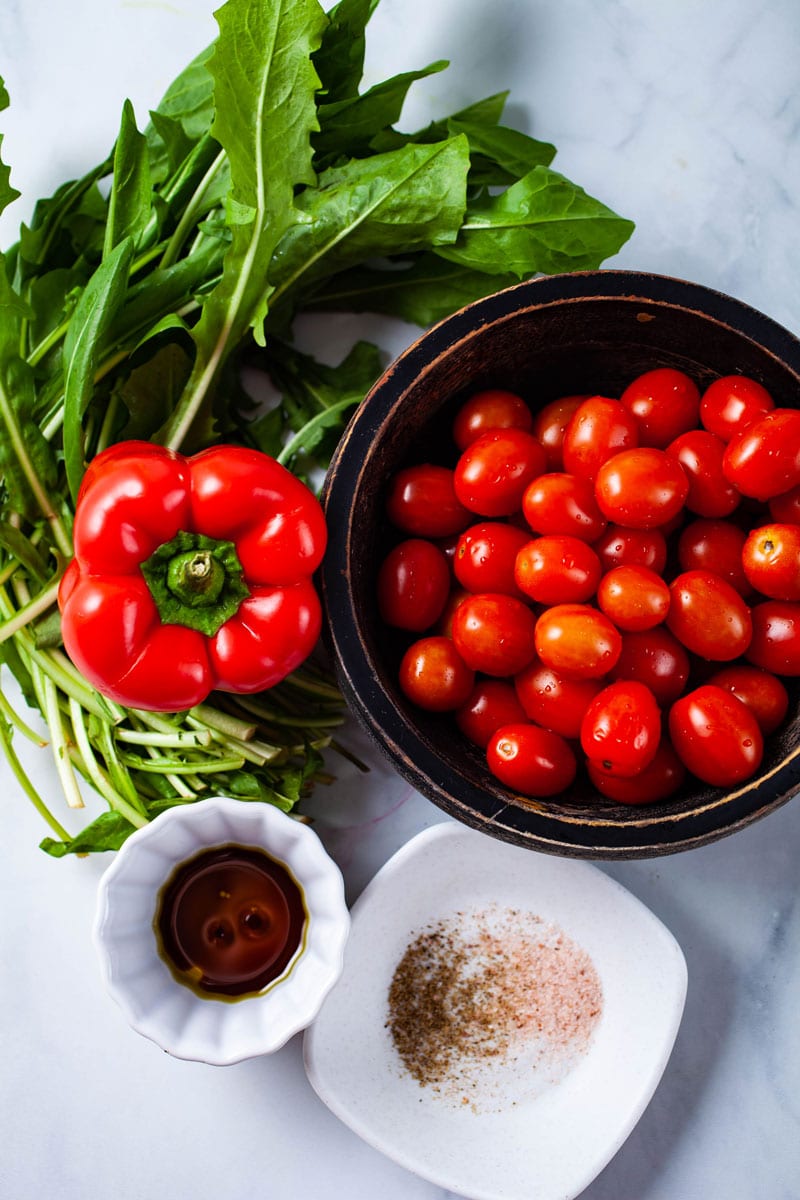 Dandelion greens, red bell pepper, small tomatoes, olive oil, balsamic vinegar, and a set of spices displayed on a table.