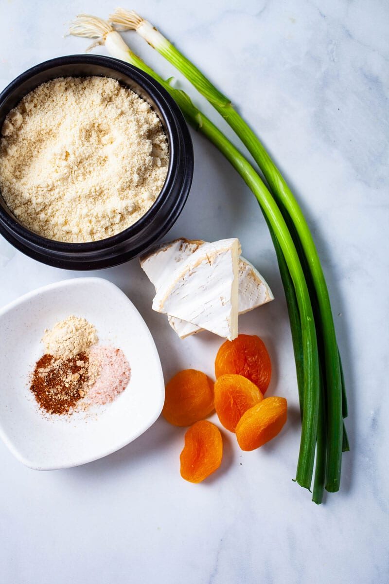 Green onions, apricots, Brie cheese, almond flour, and a set of spices displayed on a table.