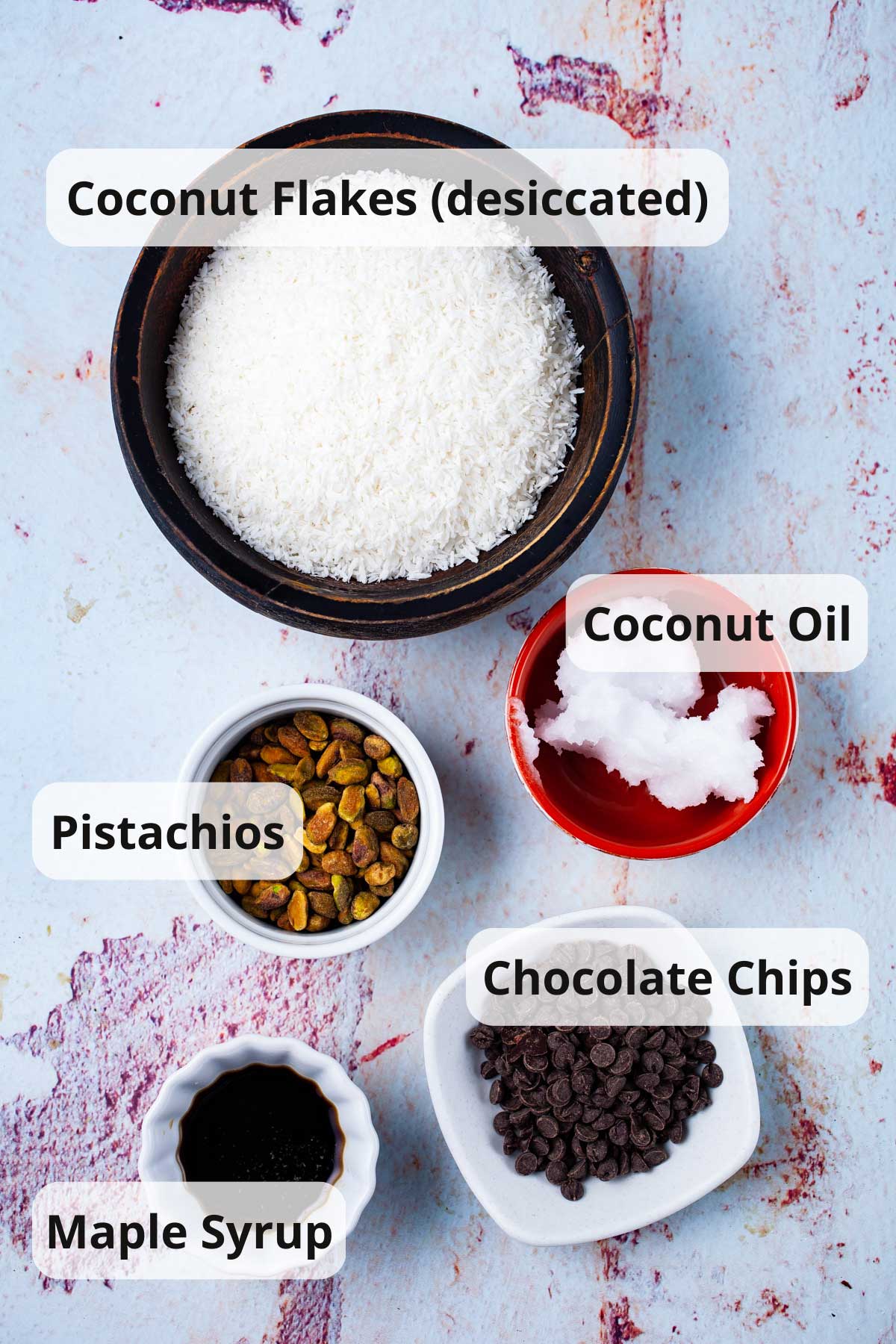 Coconut flakes, coconut oil, pistachios, chocolate chips, and maple syrup displayed on table.