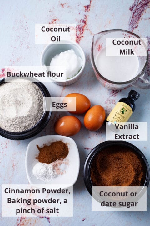 Ingredients like coconut oil, coconut milk, buckwheat flour, eggs, vanilla extract, coconut sugar, cinnamon powder, baking powder, and salt displayed on a table.