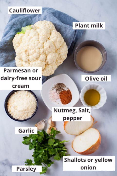 Ingredients like cauliflower, plant milk, olive oil, parmesan cheese, nutmeg, salt, pepper, garlic, parsley, and half of a yellow onion displayed on a table.