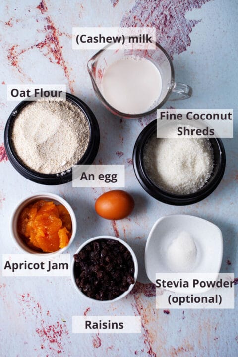 Ingredients like cashew milk, oat flour, fine coconut shreds, an egg, apricot jam, raisins, and stevia powder displayed on a table.