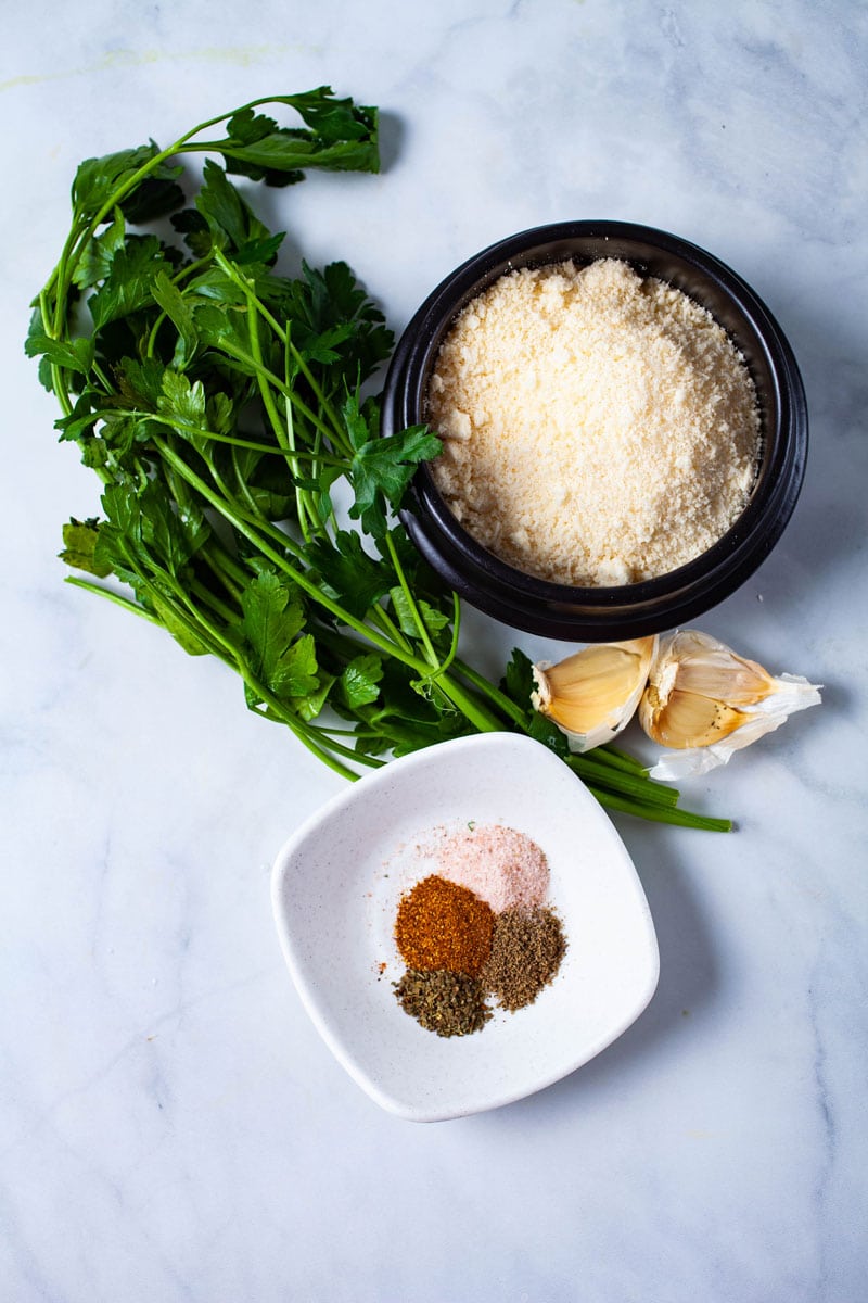 Parsley leaves, parmesan cheese, garlic cloves, and a set of spices displayed on a table.