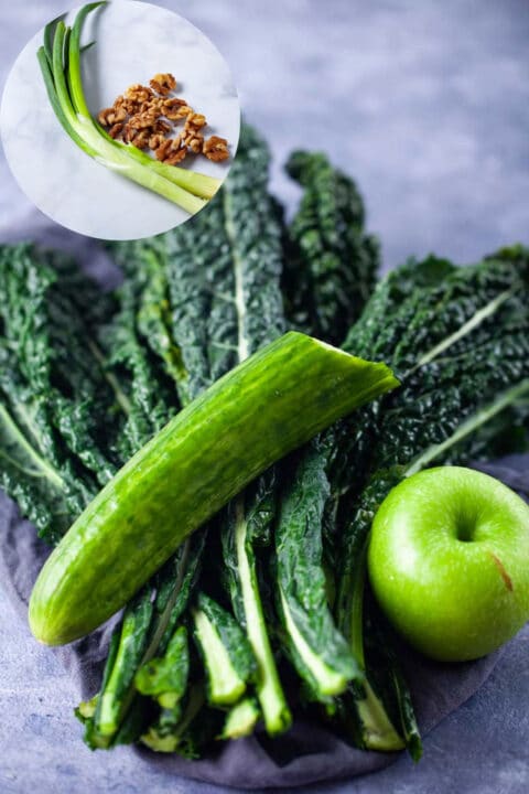 Fresk kale leaves, half a cucumber, a green apple, green onions, and chopped walnuts shown on a white background.