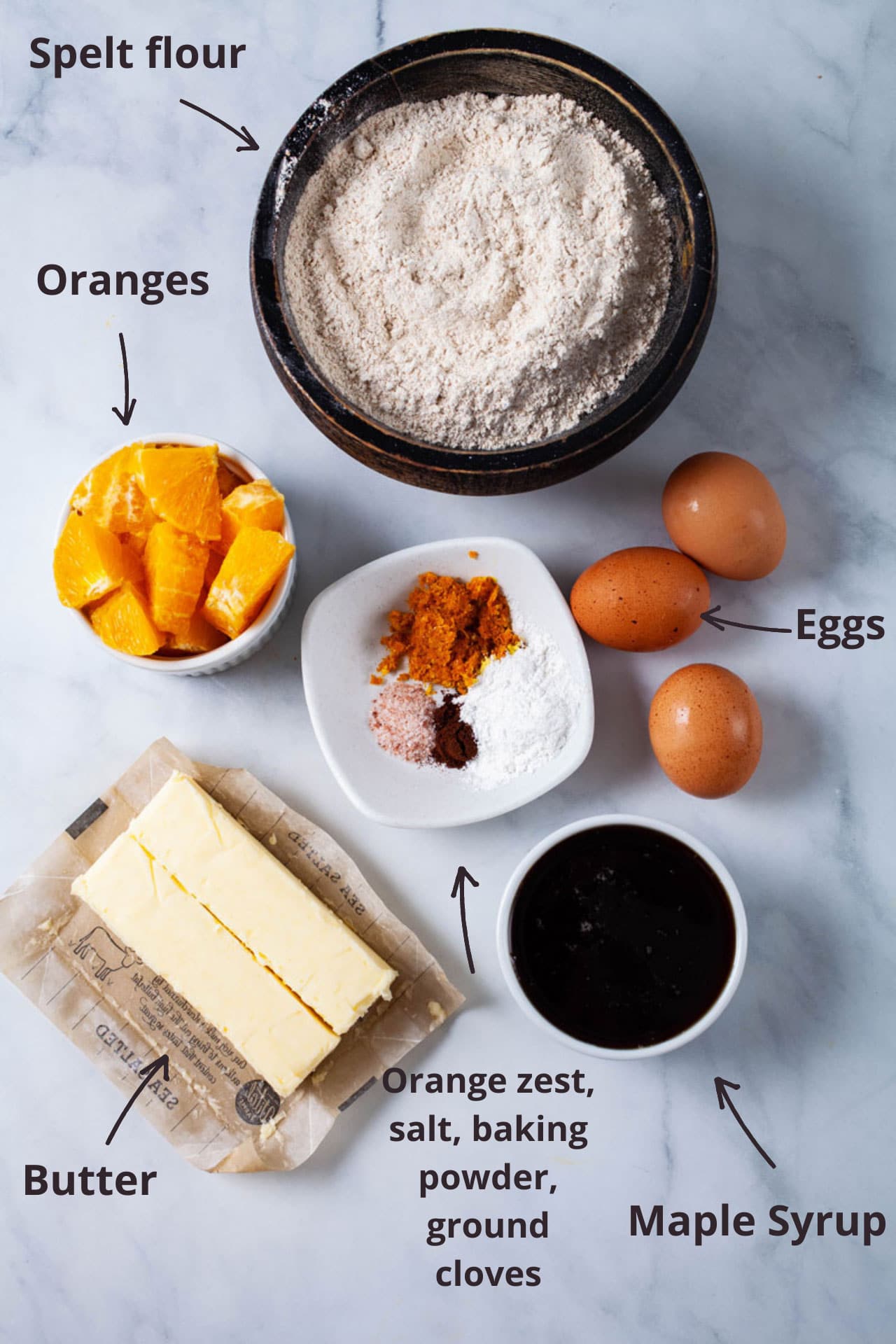 Ingredients like spelt flour, diced oranges, eggs, butter, maple syrup, orange zest, and spices displayed on a table.