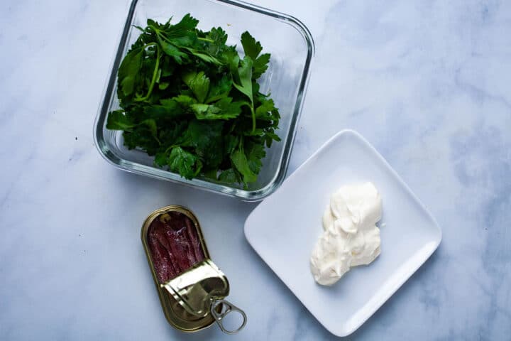 An opened can of anchovies, fresh parsley, and sour cream displayed on a table.
