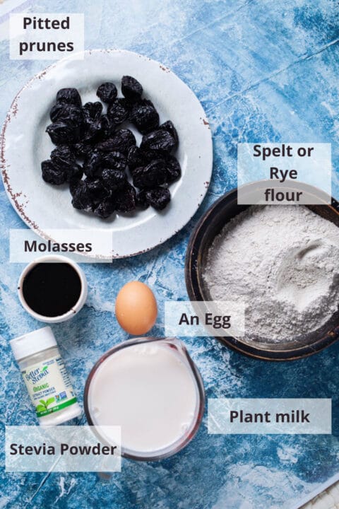 Ingredients like dried prunes, Spelt flour, Molasses, an egg, plant milk, and stevia powder displayed on a table.