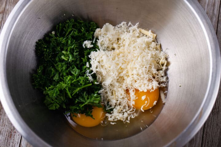 A close up of fresh chopped parsley, grated parmesan cheese, and two cracked eggs in bowl.