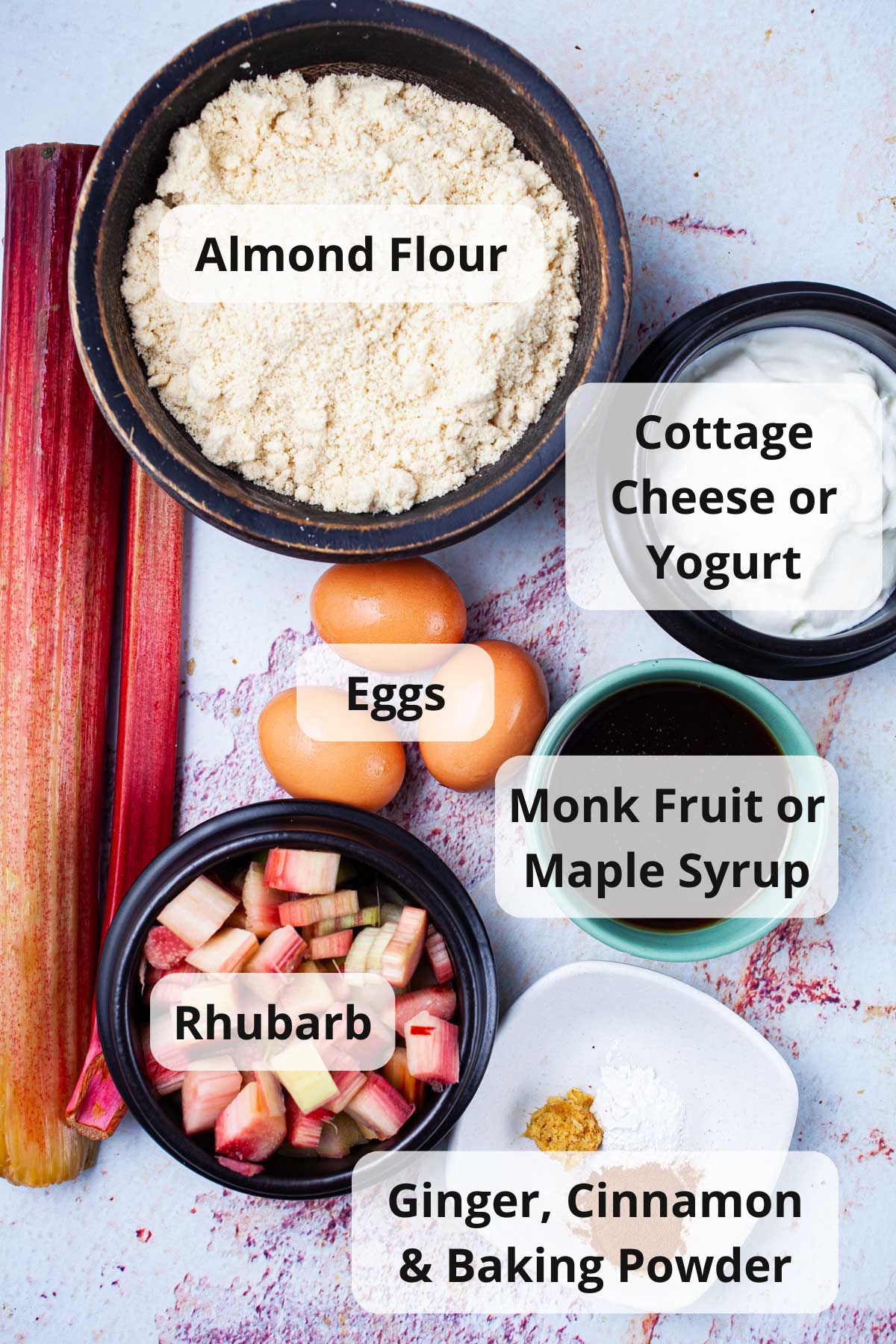 Almond flour, yogurt, eggs, maple syrup, rhubarb, ginger, cinnamon, and baking powder displayed on a table.