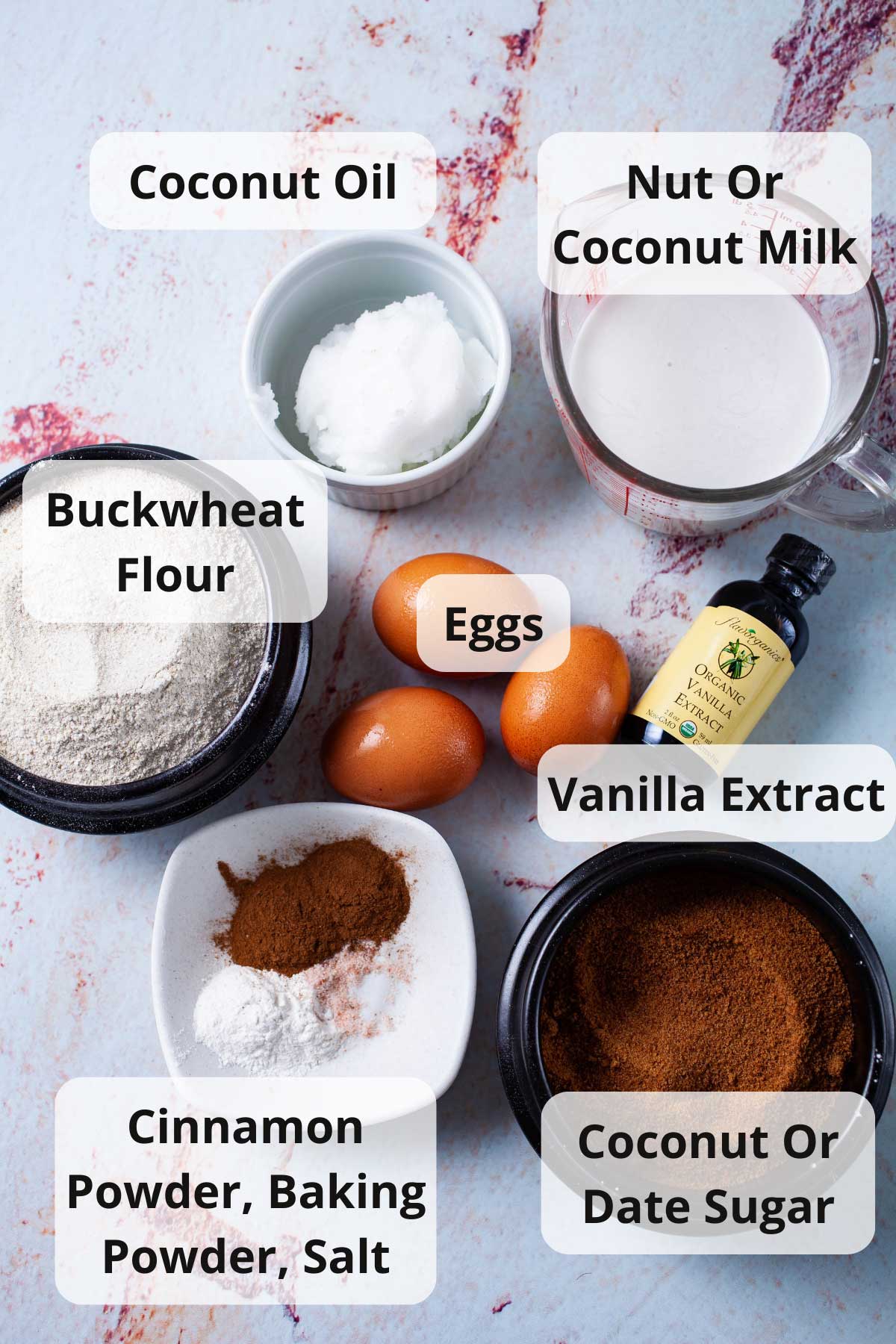 Coconut oil, coconut milk, buckwheat flour, eggs, vanilla extract, coconut sugar, cinnamon powder, baking powder, and salt displayed on a table.