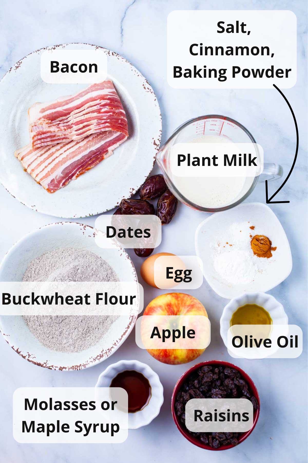 Bacon, plant milk, dates, egg, flour, apple, olive oil, molasses, raisins, baking powder, salt, and cinnamon powder displayed on a table.