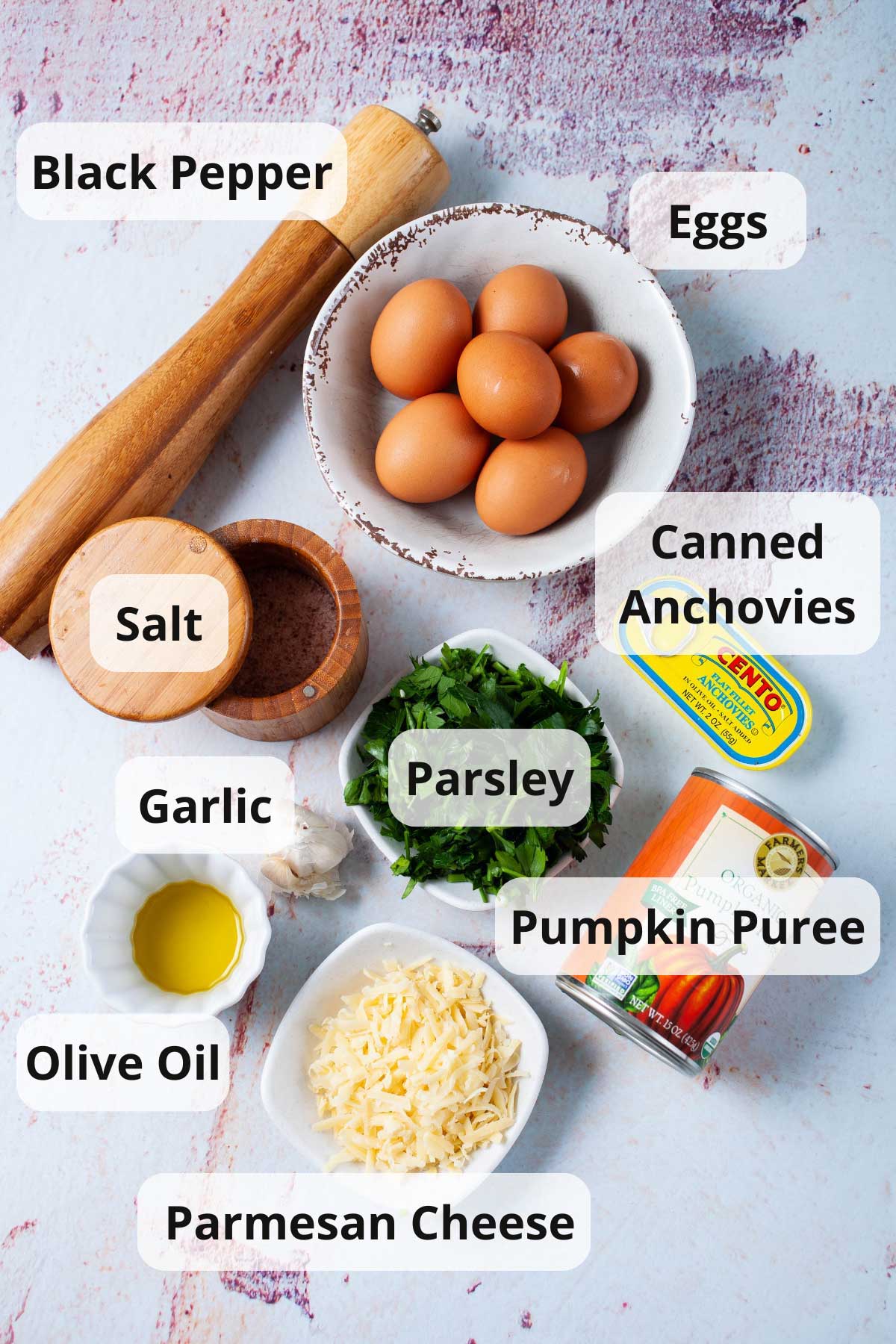 Eggs, black pepper, salt, garlic, olive oil, Parmesan cheese, parsley, pumpkin puree, and canned anchovies displayed on a table.