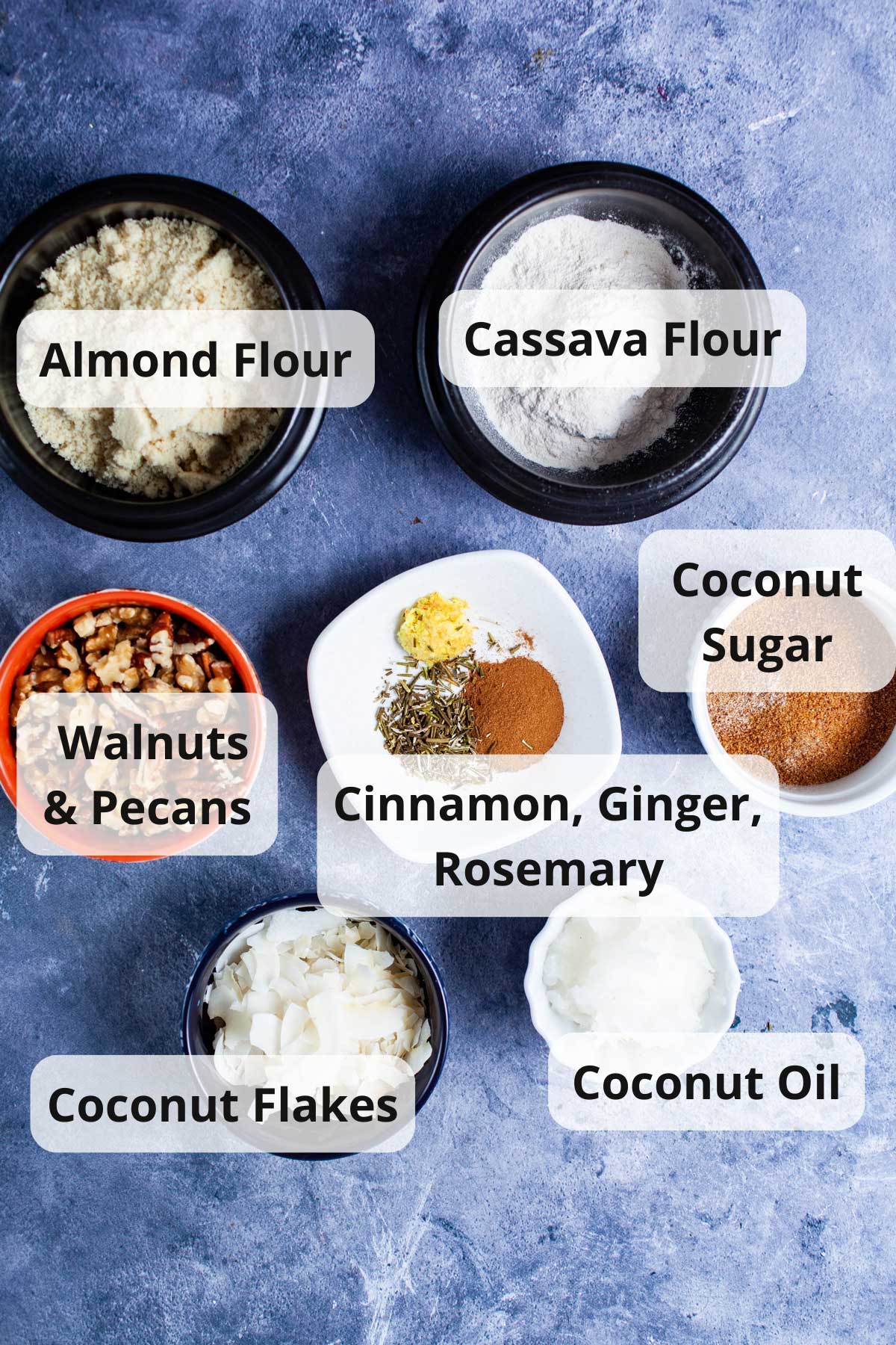 Coconut oil, coconut flakes, chopped walnut & pecans, almond flour, cinnamon, ginger, dried Rosemary, Cassava flour, coconut sugar displayed on a table.