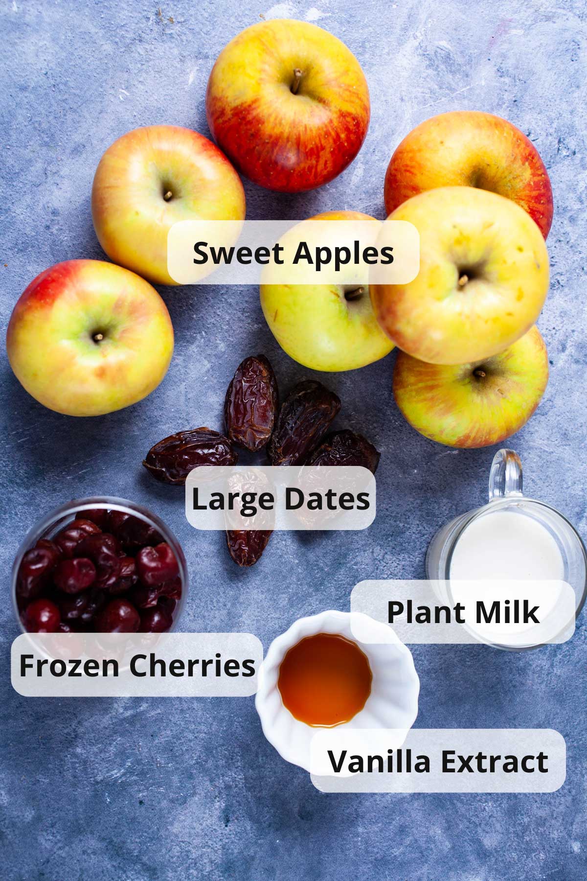 Sweet apples, large dates, plant milk, frozen cherries, plant milk, and vanilla extract displayed on a table.