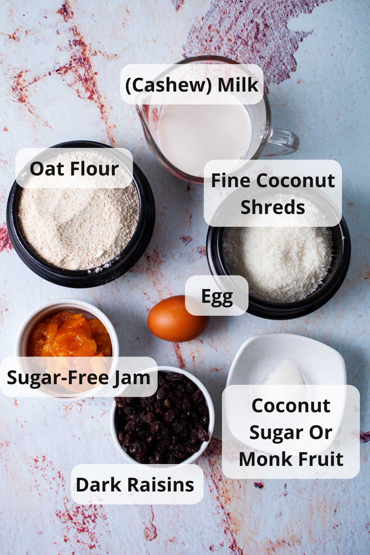 Cashew milk, oat flour, fine coconut shreds, egg, sugar, raisins, and apricot jam displayed on a table.