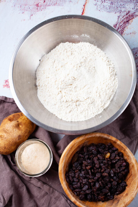 Ingredients such as a bowl of flour, a russet potato, leavened yeast, and a bowl of raisins displayed on a table.