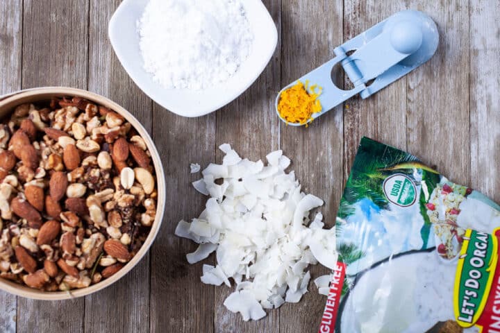 A bowl of mixed nuts, powdered sugar, orange zest, and coconut shreds displayed on a wooden board.