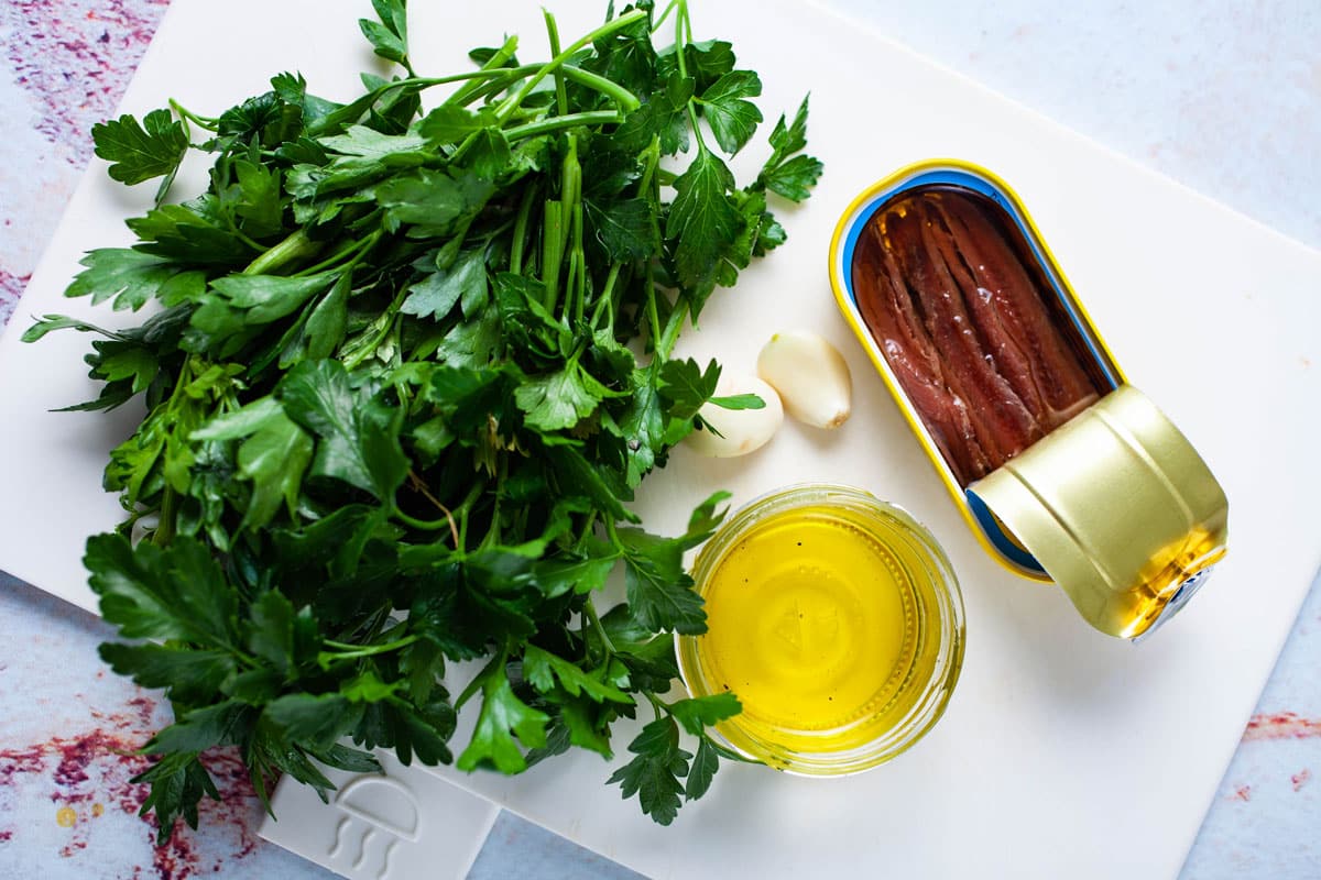Ingredients like canned anchovies, fresh Parsley, olive oil, and garlic cloves displayed on a white cutting board.