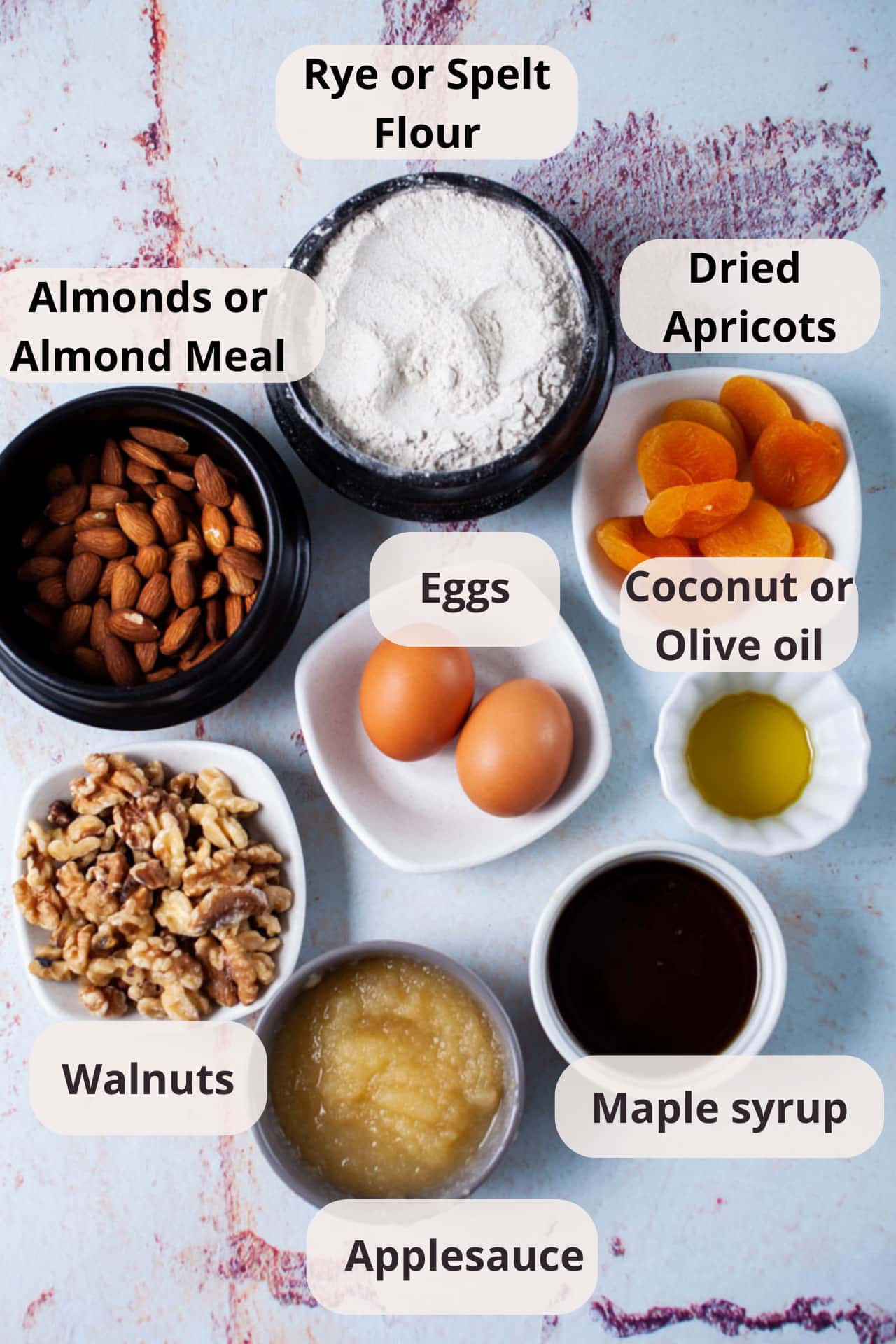 Rye flour, dried apricots, olive oil, maple syrup, applesauce, walnuts, eggs, almonds displayed in separate bowls on a table.