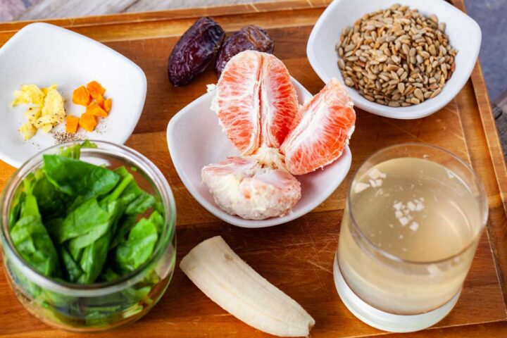 A wooden board displaying several ingredients like seeds, banana, orange, turmeric, ginger, coconut water, spinach, and dates to make a green smoothie.