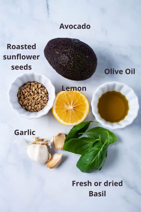 Ingredients such as avocado, roasted sunflower seeds, olive oil, lemon, garlic, and fresh basil laid out on a board.