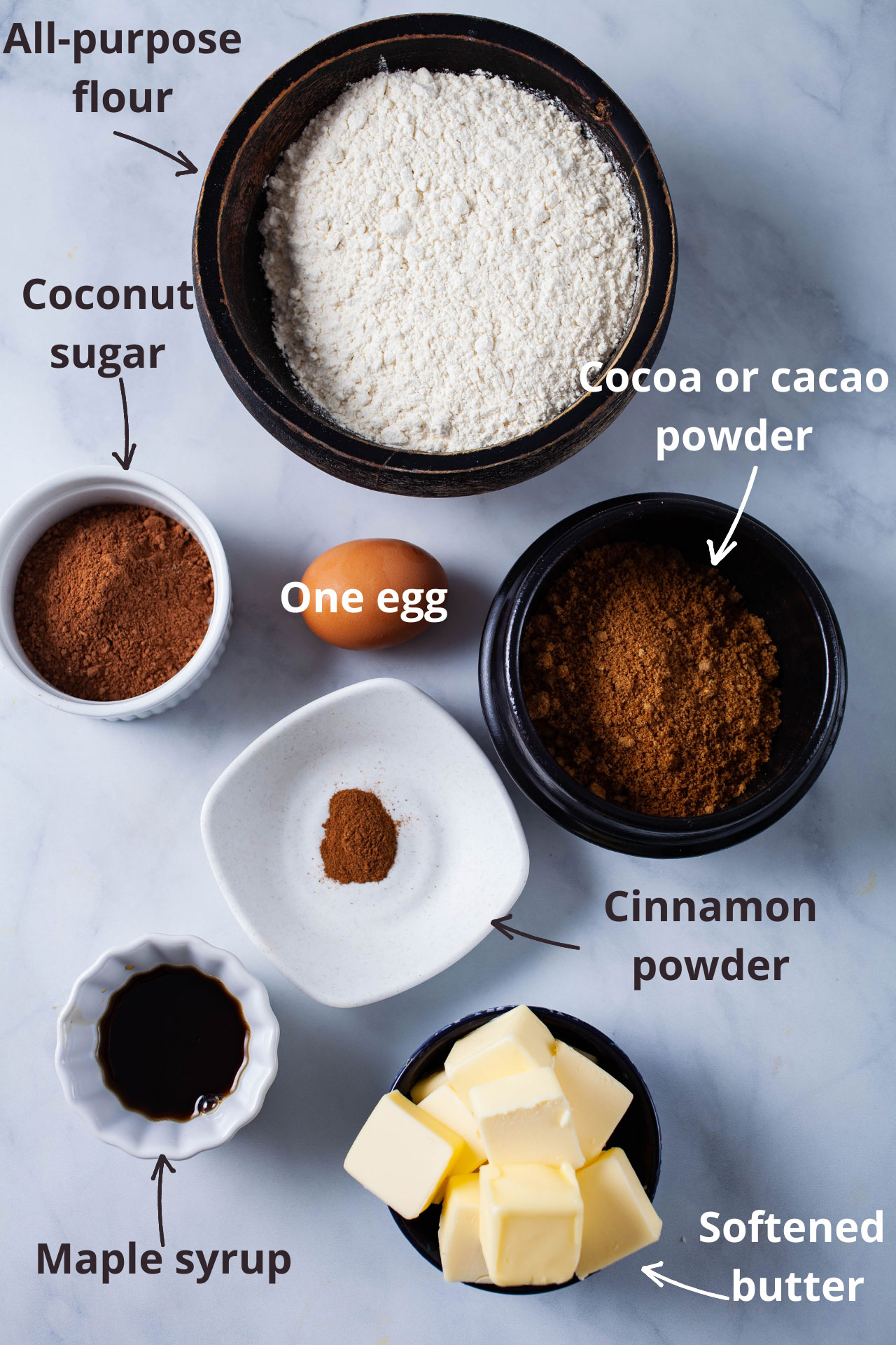 Ingredients like all-purpose flour, coconut sugar, cacao powder, an egg, cinnamon powder, maple syrup, and softened butter displayed on a table.