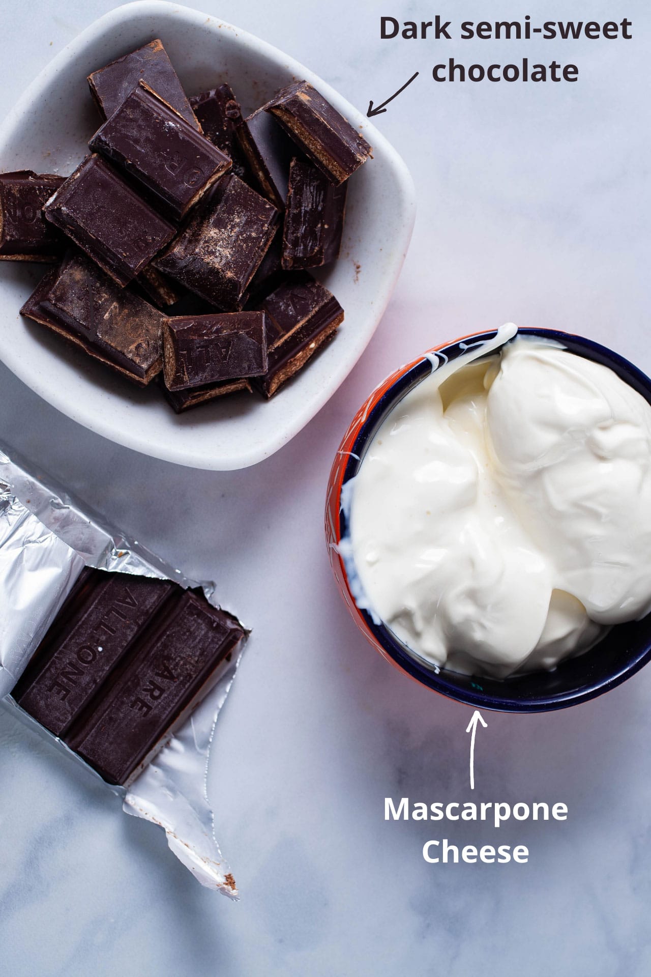 Ingredients like dark chocolate and a bowl of mascarpone cheese displayed on a table.