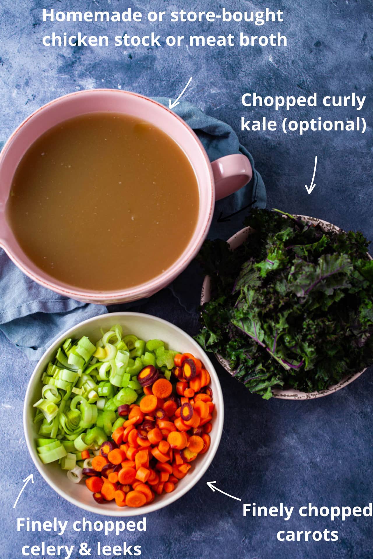 A large bowl with chicken stock, another bowl with chopped fresh veggies like carrots, leeks, and celery, and another bowl with chopped kale on a table.