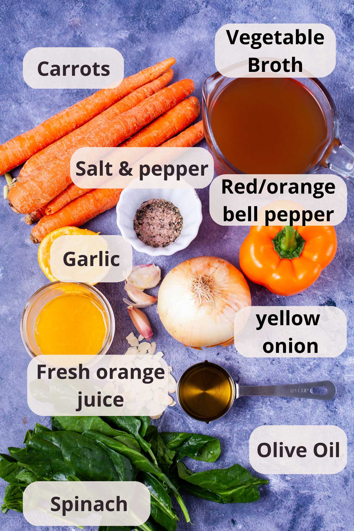 Ingredients like vegetable broth, olive oil, salt and pepper, garlic, yellow onion, carrots, shaved almonds, orange bell pepper, spinach leaves, and fresh orange juice displayed on a table.