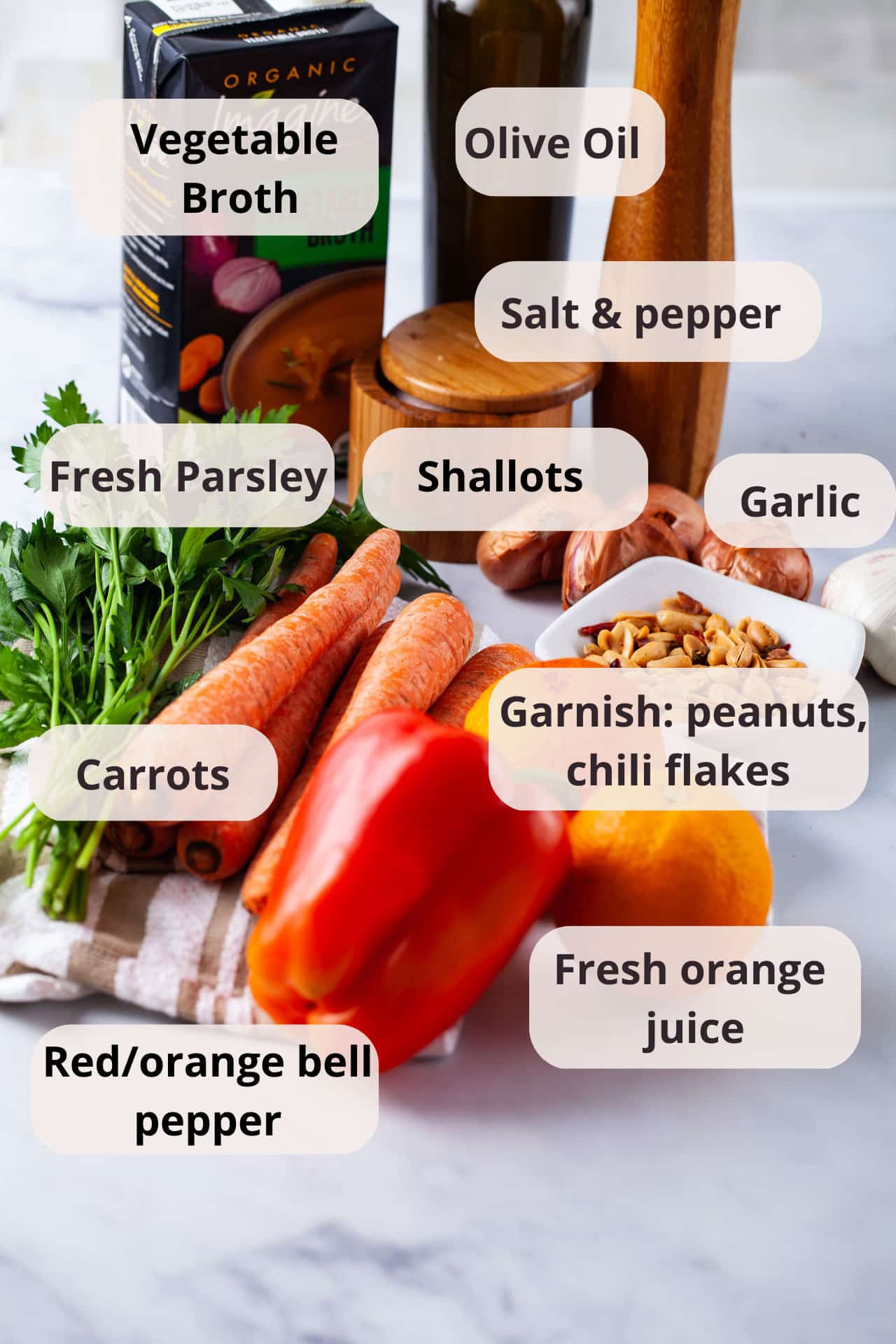 Ingredients like vegetable broth, olive oil, salt and pepper, garlic, shallots, carrots, spicy peanuts, red bell pepper, and fresh oranges displayed on a table.