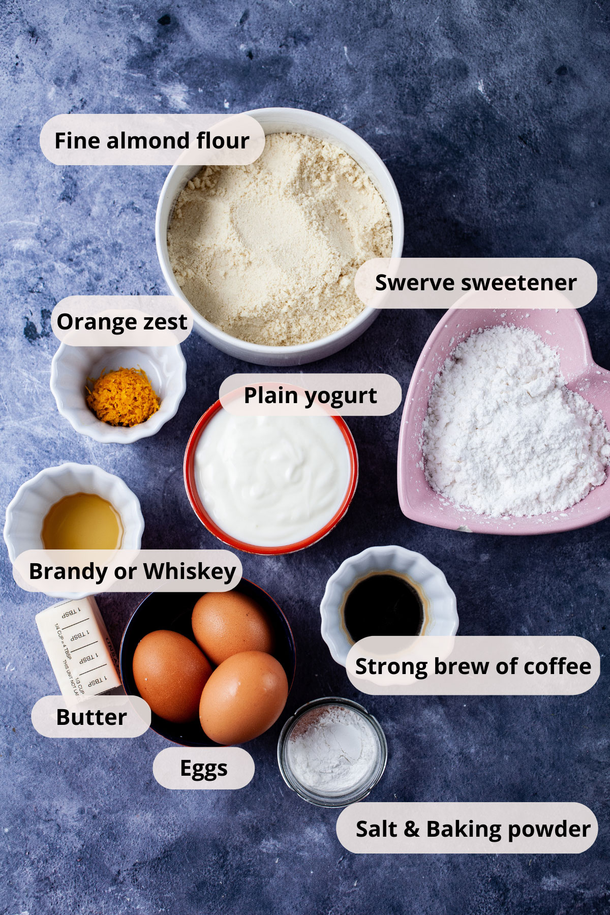 Ingredients like almond flour, Swerve, orange zest, vanilla extract, yogurt, 3 eggs, butter, Brandy, brewed coffee, salt and baking powder displayed on a table in individual bowls.