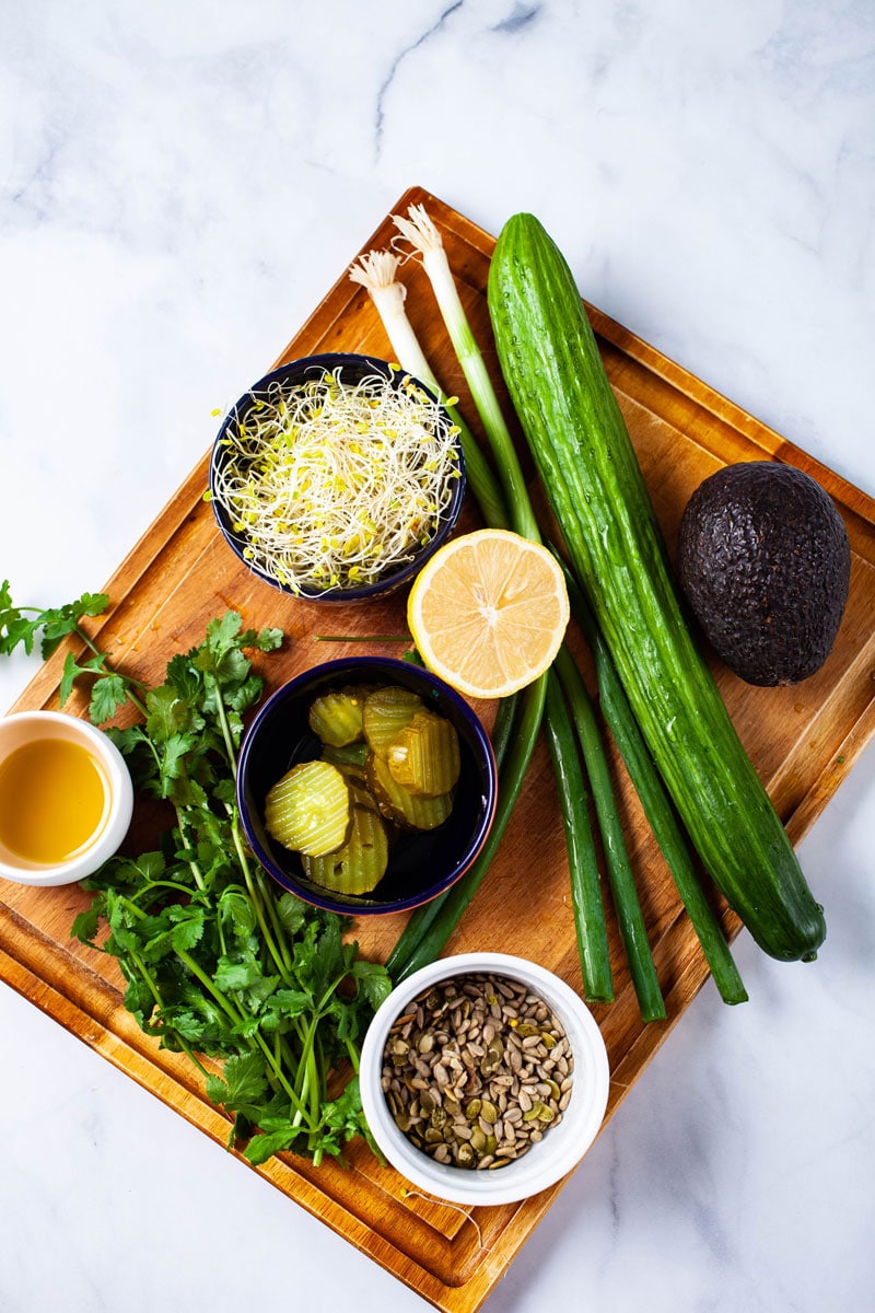 A wooden board serving olive oil, sweet pickles, cucumber, green onions, lemon, avocado, pumpkin seeds, cilantro leaves, and broccoli sprouts to prepare a salad.