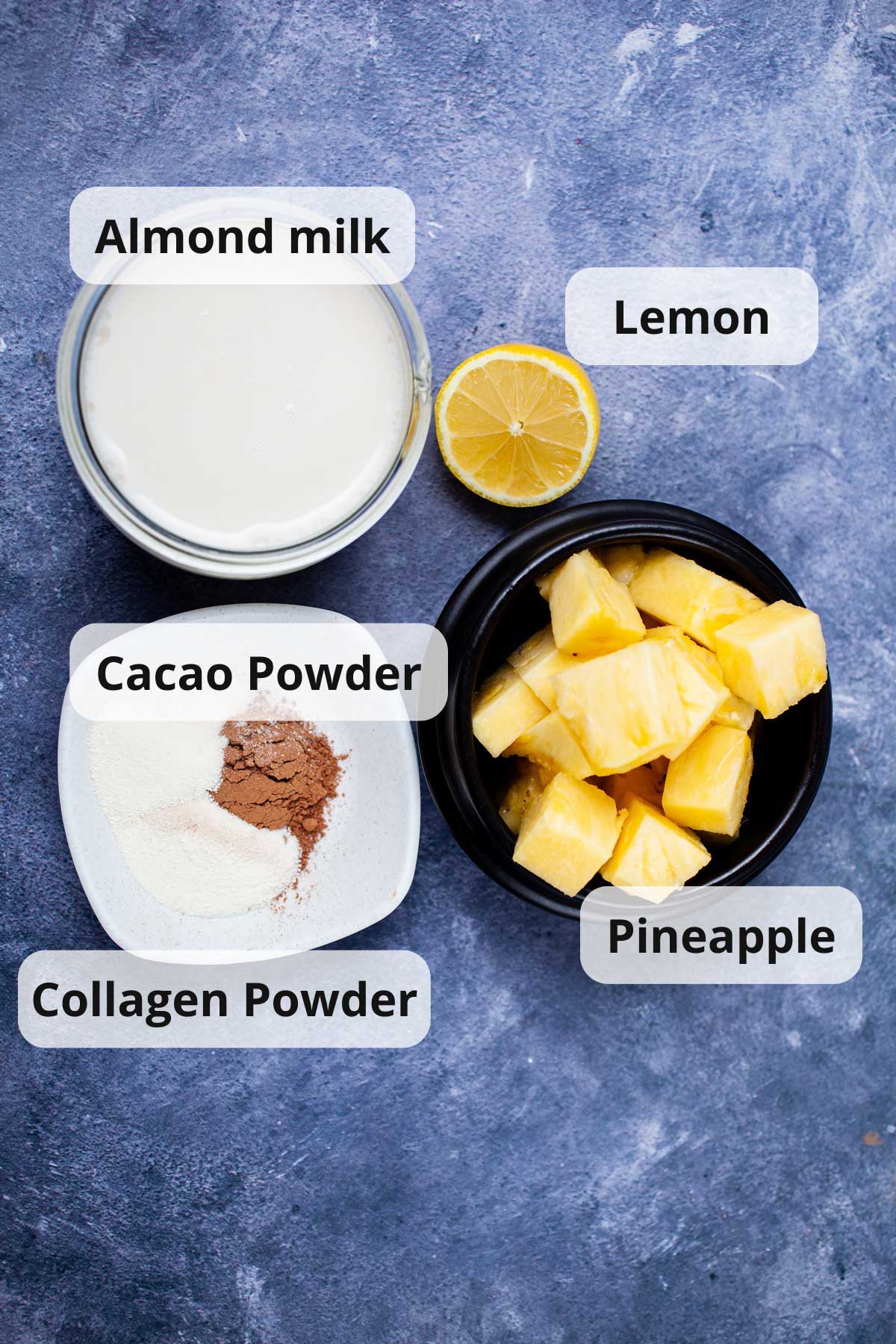 Fresh chopped pineapple, half of a lemon, almond milk, cacao powder and collagen powder displayed on a table.