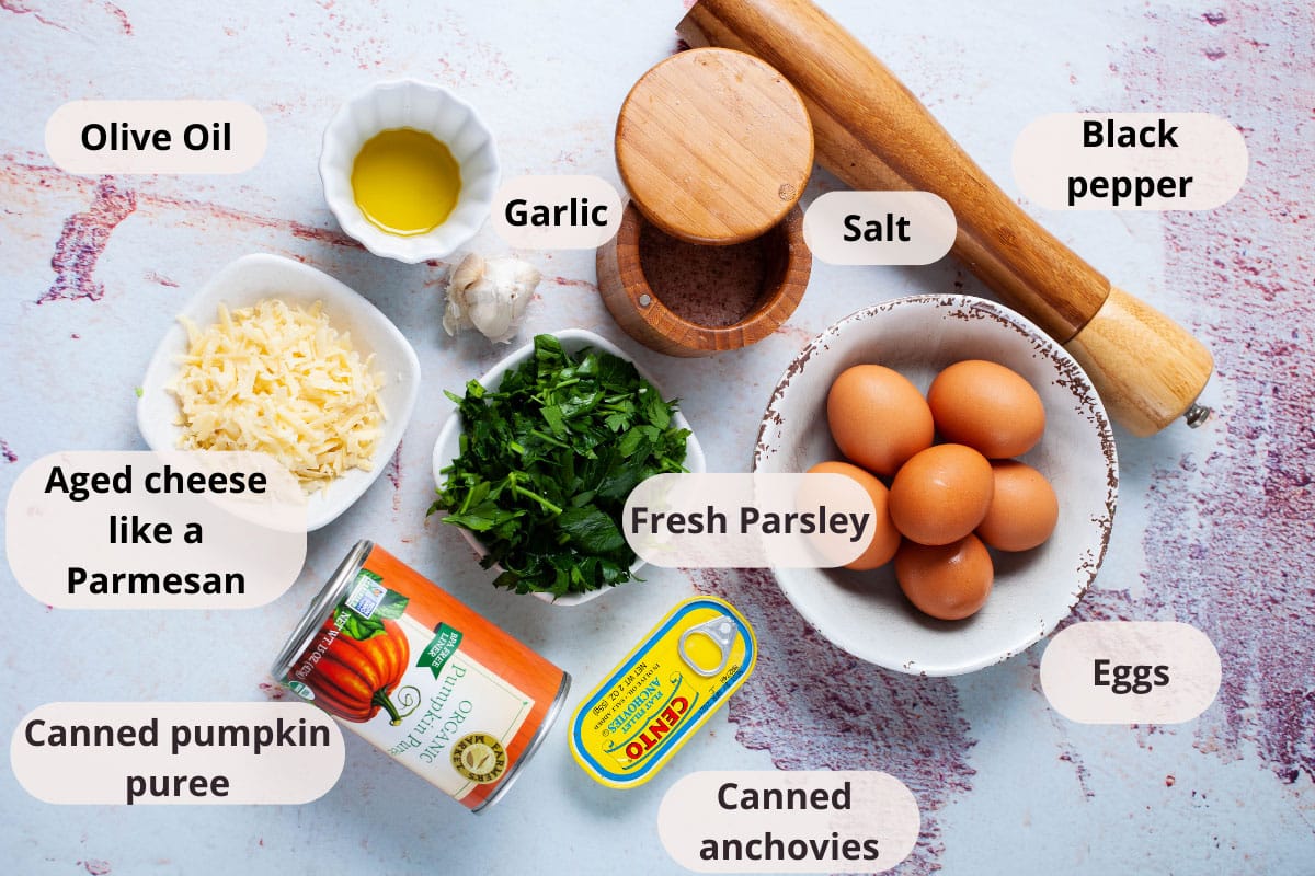 Olive oil, garlic, salt, black pepper mill, eggs, fresh parsley, canned anchovies, canned pumpkin puree, and grated Parmesan cheese displayed on a table.