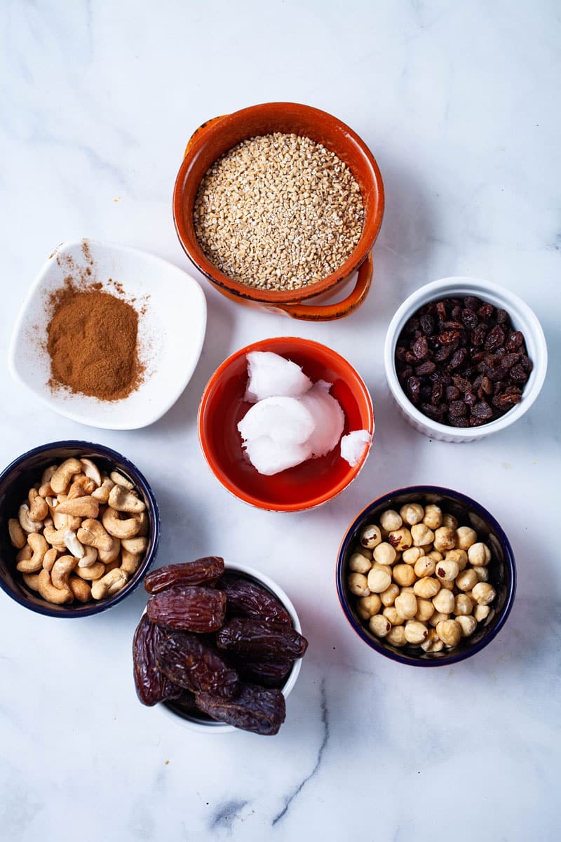 Oats, cinnamon, coconut oil, raisins, cashews, dates, and hazelnuts displayed on a table.