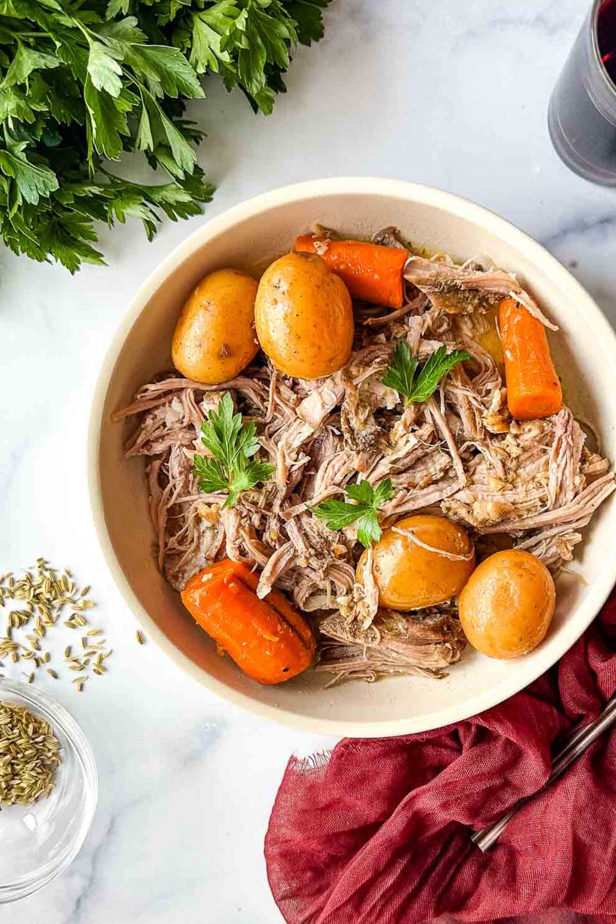 Pork roast in a bowl with small potatoes and carrots, topped with parsley leaves.