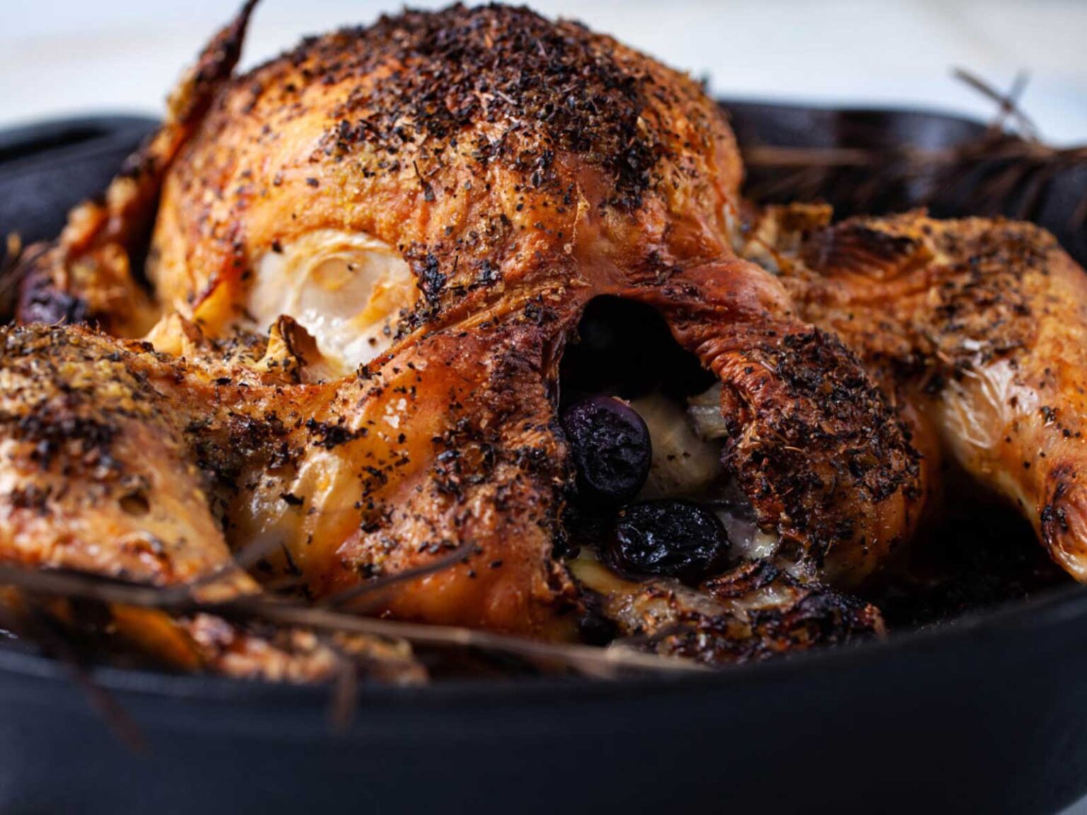A roasted chicken is served in a black dish. The chicken is golden brown and seasoned with herbs, and its cavity is stuffed with what appears to be fruits and vegetables, including dark purple olives.