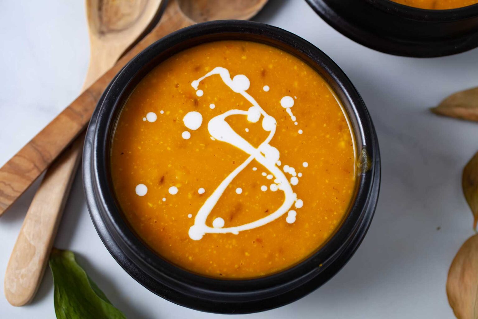 A black bowl filled with creamy orange soup, garnished with a swirl of white cream. Two wooden spoons and a green leafy garnish are placed beside the bowl on a white surface. Another black bowl is partially visible in the background.