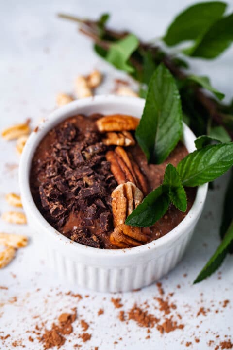A ramekin filled with chocolate chia pudding topped with shaved chocolate, pecans, and fresh chocolate mint leaves.