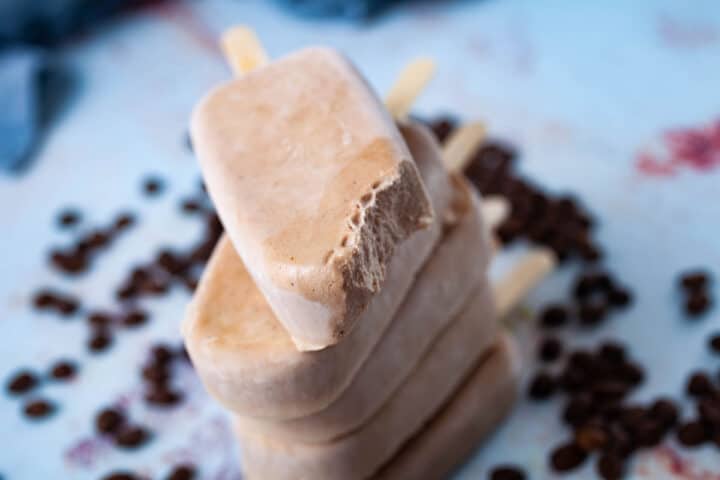 A bite out of a brown colored ice pop, stacked on top of other ice pops and surrounded by coffee beans.