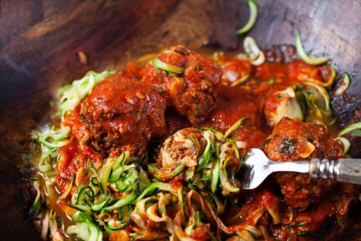 A large wooden bowl filled with raw zucchini noodles topped with an Italian red spaghetti sauce and beef meatballs.