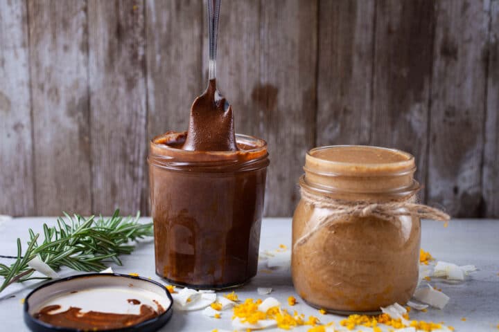 A small spoon lifting creamy chocolate nut butter out of a jar and next to another jar with coconut butter.
