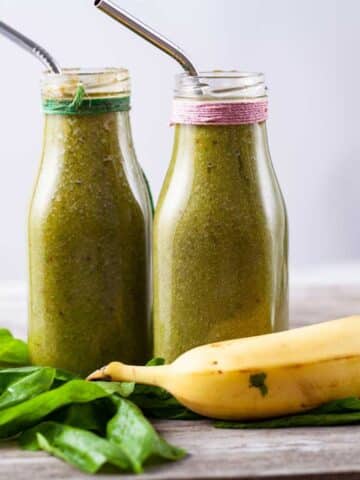 Two milk bottles filled with green smoothie drink, surrounded by half a banana and fresh spinach.