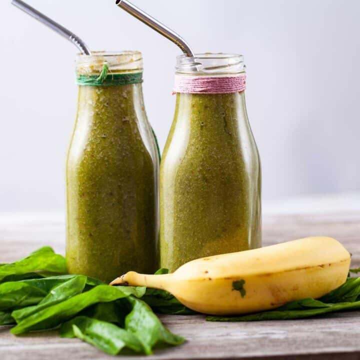 Two milk bottles filled with green smoothie drink, surrounded by half a banana and fresh spinach.