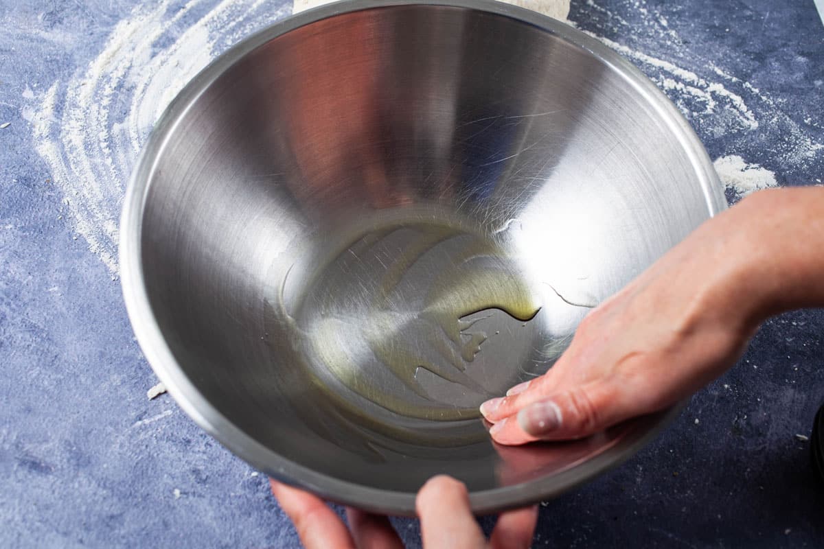 A hand coating a stainless steel bowl with olive oil.
