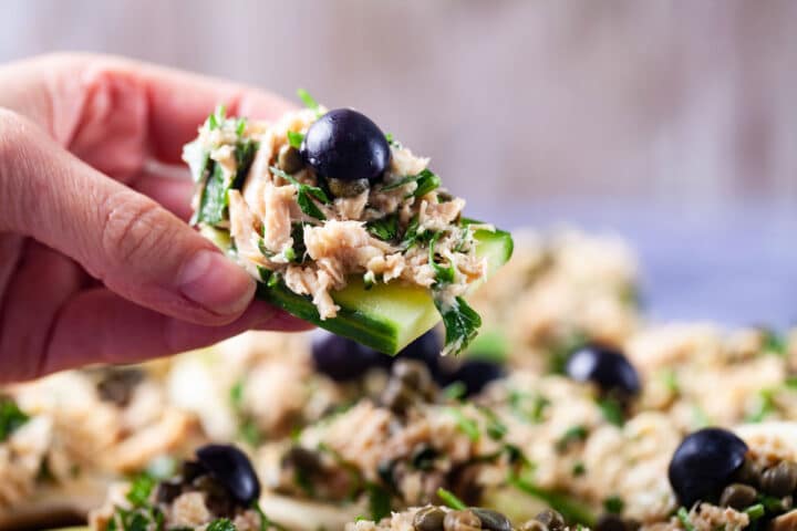 A hand holding a thick cucumber slice topped with creamy tuna and parsley salad and garnished with dark grapes.