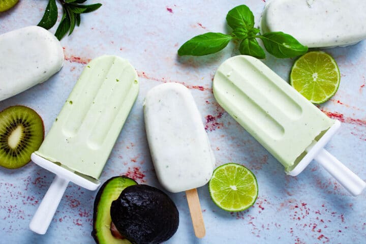 White and green ice pops displayed on a table and surrounded by slices of lime, kiwi, avocado, and mint leaves.